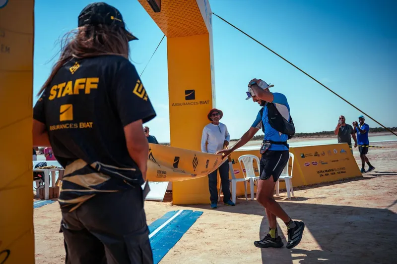 The Ultra Mirage El Djerid finish line: a runner crosses the tape while Adel Beznine stands by the arch.
