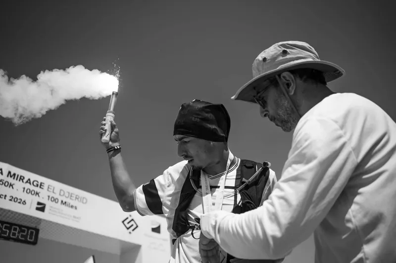 A runner holds up a smoke flare at the finish while Adel Beznine checks his watch.